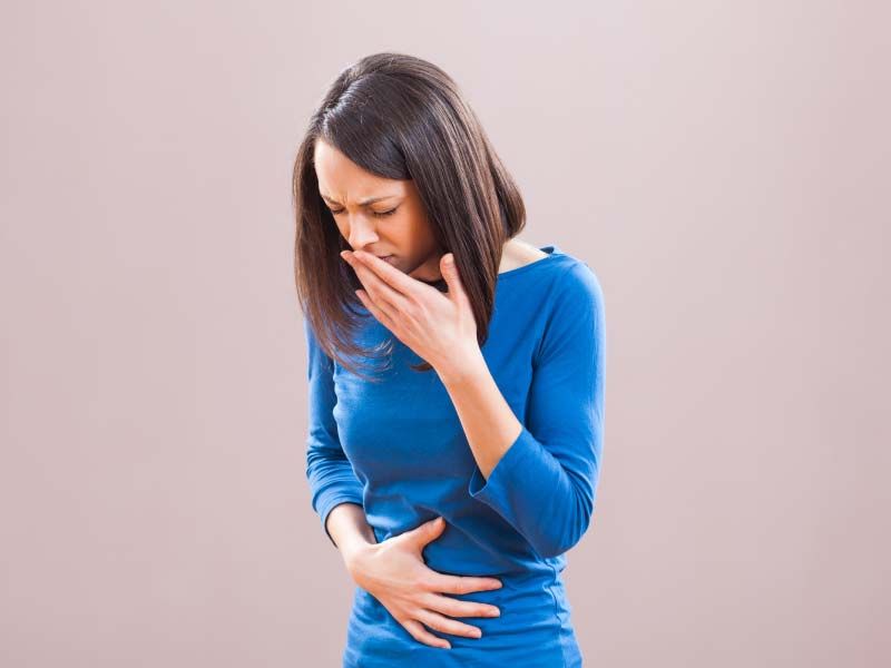 A woman wearing a blue shirt has one hand over her abdomen and one over her mouth indicating nausea