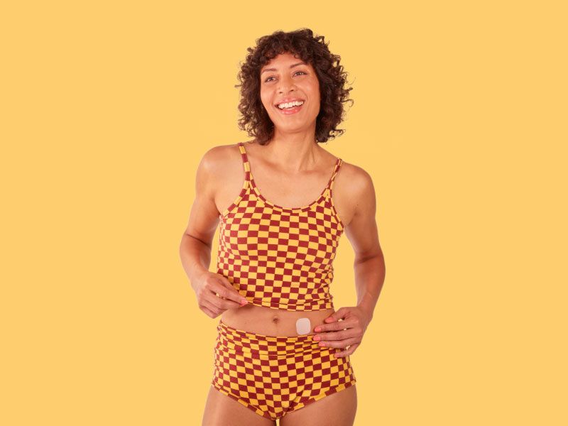 A woman smiling and wearing a checkered yellow and brown tank top and shorts is placing a hormone replacement therapy patch on her abdomen
