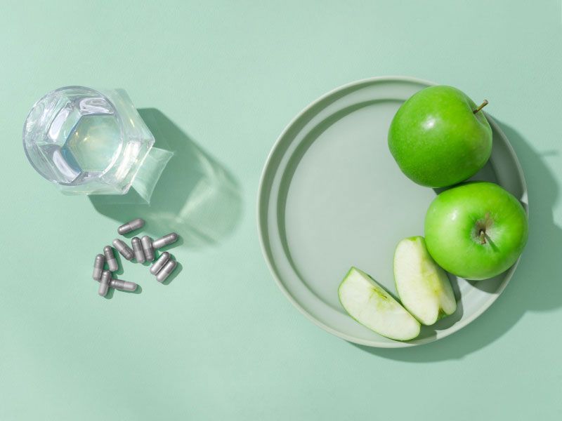Sliced and whole apples on a plate with supplement capsules next to a glass of water on a green surface