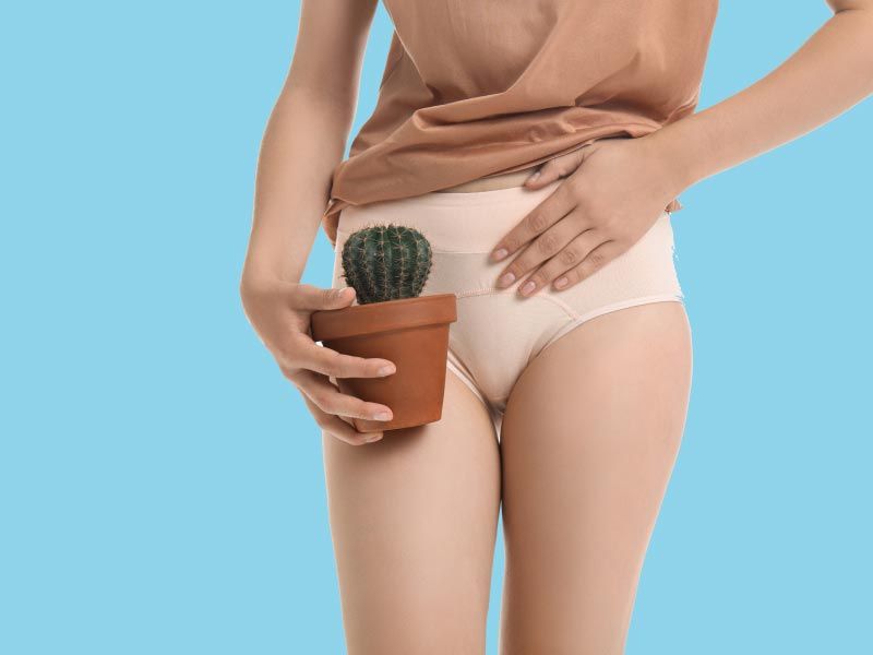 A woman standing up in white underwear and a tan shirt with a hand on her abdomen and holding a small cactus indicating pain