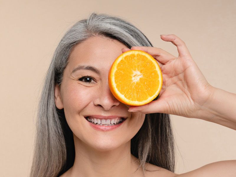 A woman is smiling and holding a sliced open orange in front of her left eye with a beige background