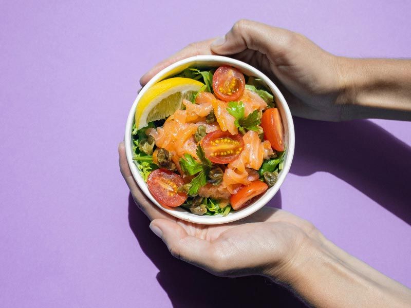A person's hands holding a bowl of fruits and veggies on a purple surface