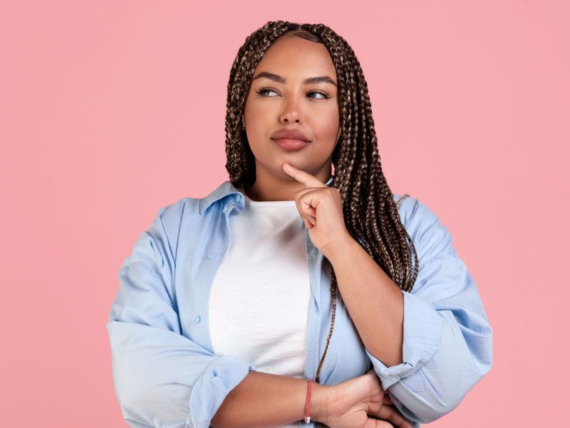 A woman wearing a white tee with a light blue button-up shirt has a finger to her chin in contemplation in front of a pink background