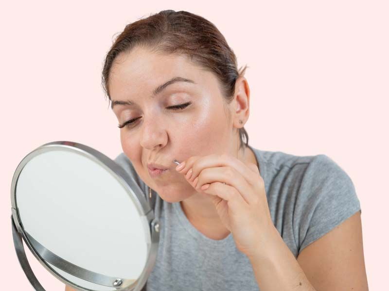 A woman wearing a gray shirt looking at her face in a hand mirror in front of a pink background