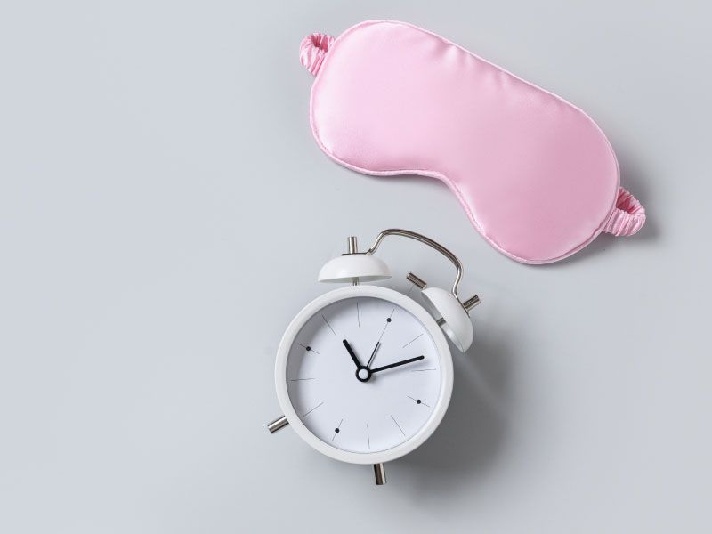 A pink sleep mask and a white alarm clock on a light gray surface