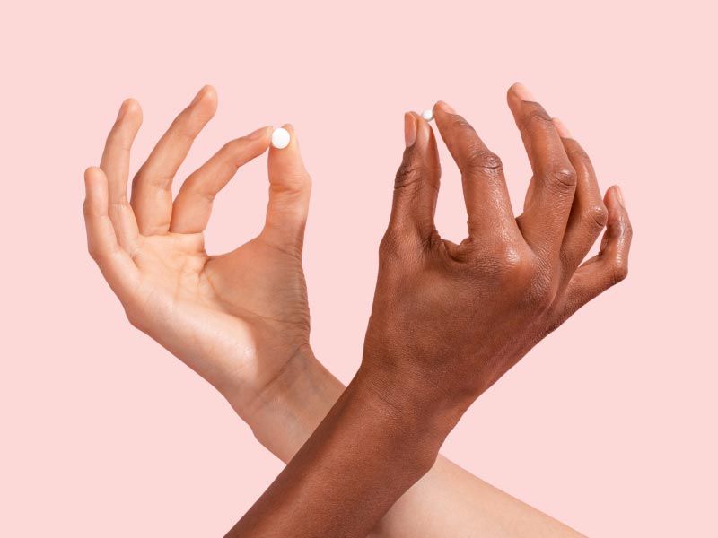 Two female hands criss crossing, both holding an Emergency Contraception pill in front of a pink background