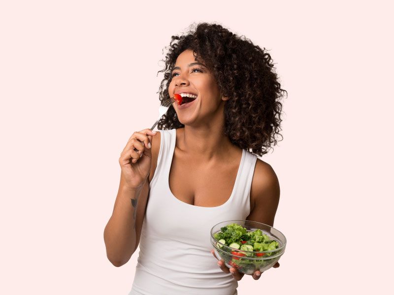 A woman wearing a white tank top is smiling while eating a salad