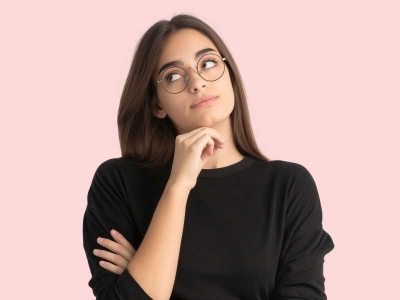 A woman wearing a black sweater has her hand on her chin in contemplation in front of a pink background