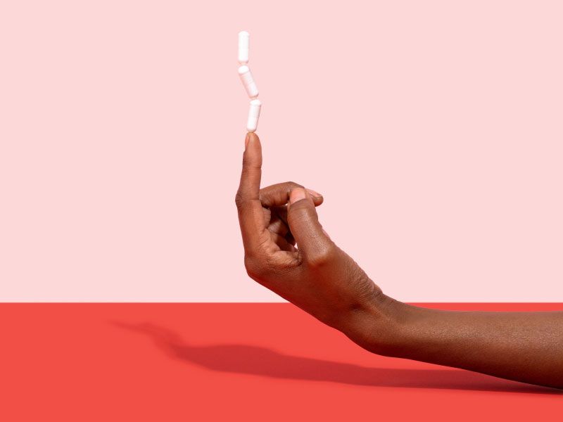 A woman's hand with 3 Boric Acid capsules balancing on her pointer finger on a red surface with a pink background