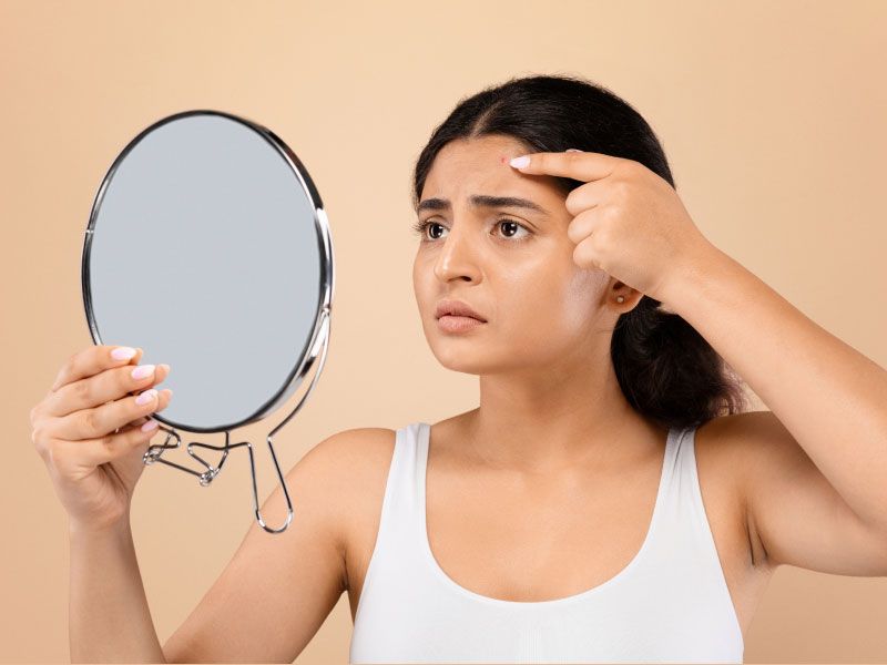 A woman wearing a white tank top is looking into the mirror while touching her forehead