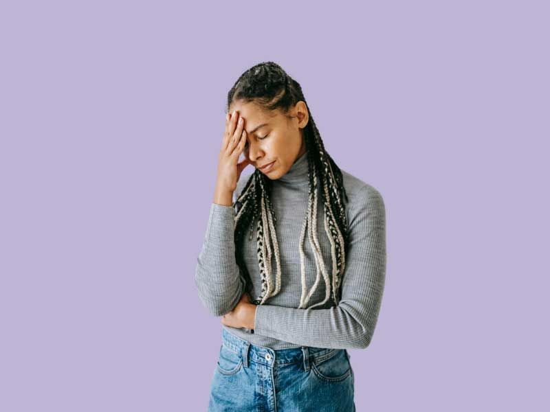 A woman wearing a gray shirt and jeans has her hand on her forehead indicating fatigue