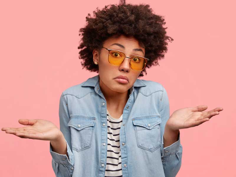 Woman wearing a striped shirt and a denim jacket is shrugging shoulders with a confused look on her face