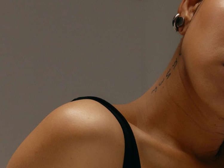 Close-up photo of a woman's shoulder and neck, wearing a black strap top, with a small script-style tattoo visible on the side of her throat.