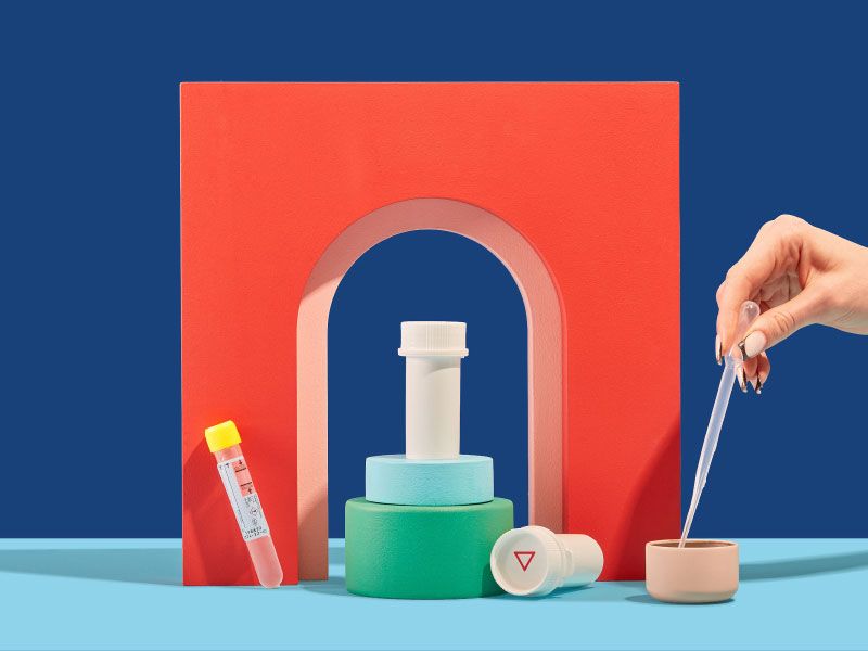 Wisp pill vials, a testing tube, and a woman's hand holding a pipette are balanced on colorful abstract shapes on a light blue surface with a dark blue background