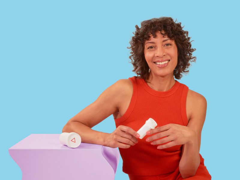 A woman wearing a red tank top is smiling while holding a Wisp pill vial next to a purple table