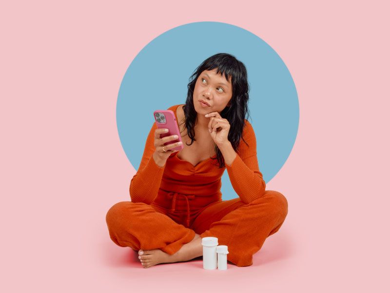 A woman wearing a red shirt and pants its sitting on a pink surface with a mobile phone and a confused look on her face