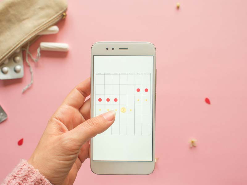 A woman's hand holding a mobile phone with a calendar tracking her menstrual cycle with tampons and menstruation medication on a pink surface in the background