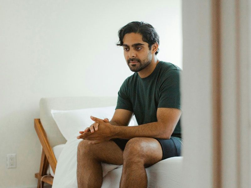 A man wearing a gray shirt and shorts is sitting on a bed with a worried look on his face