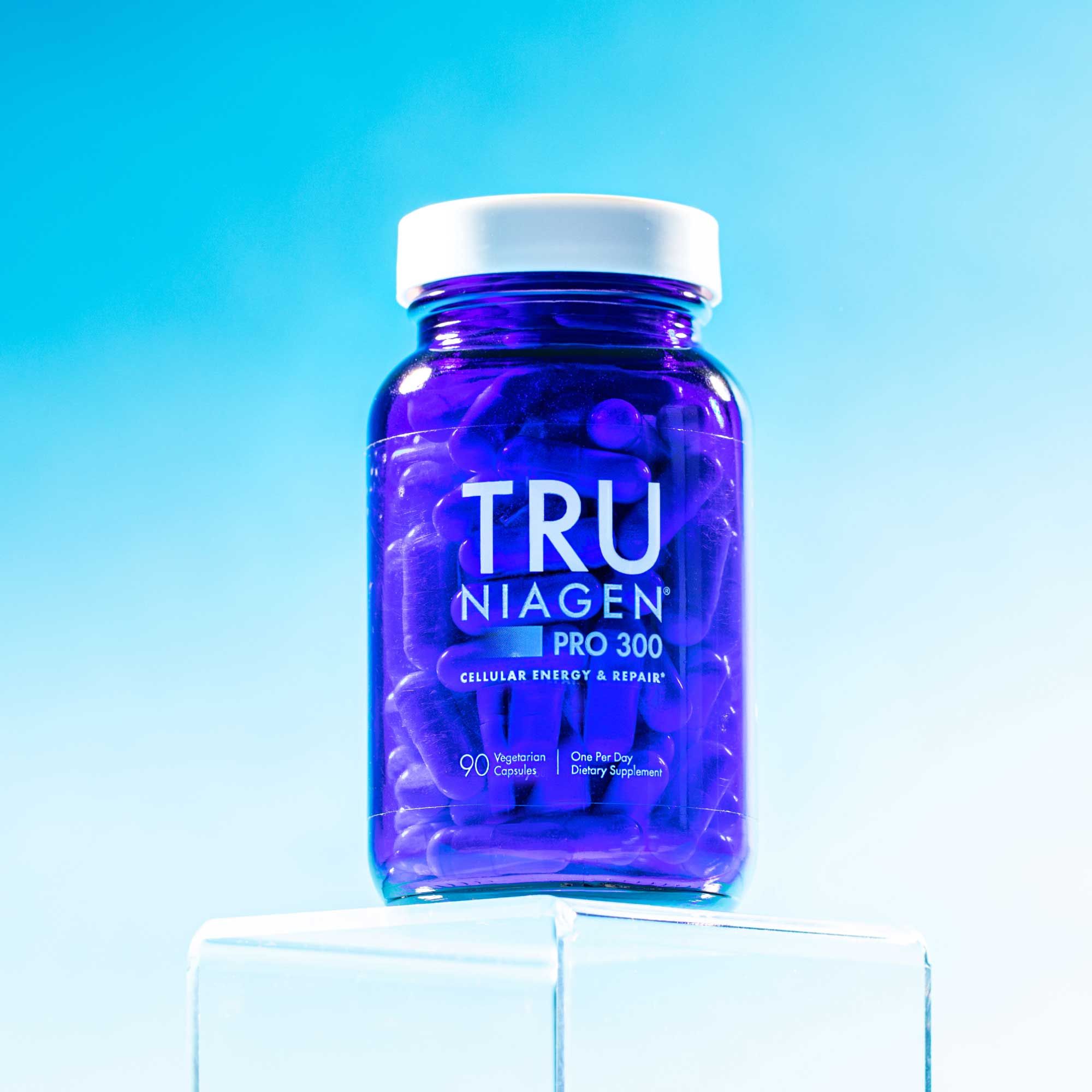 A blue Tru Niagen bottle balanced on a clear cube with a light blue gradient background