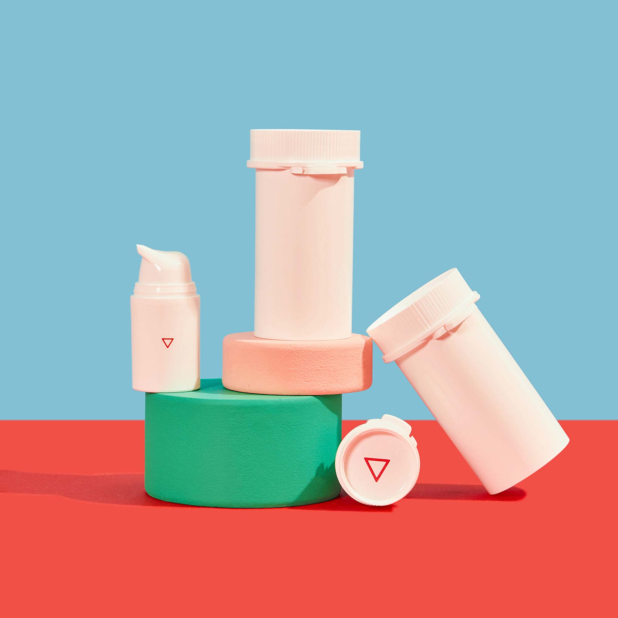 Wisp plastic pill vials and a pump bottles balanced on colorful abstract shapes on a red surface with a light blue background
