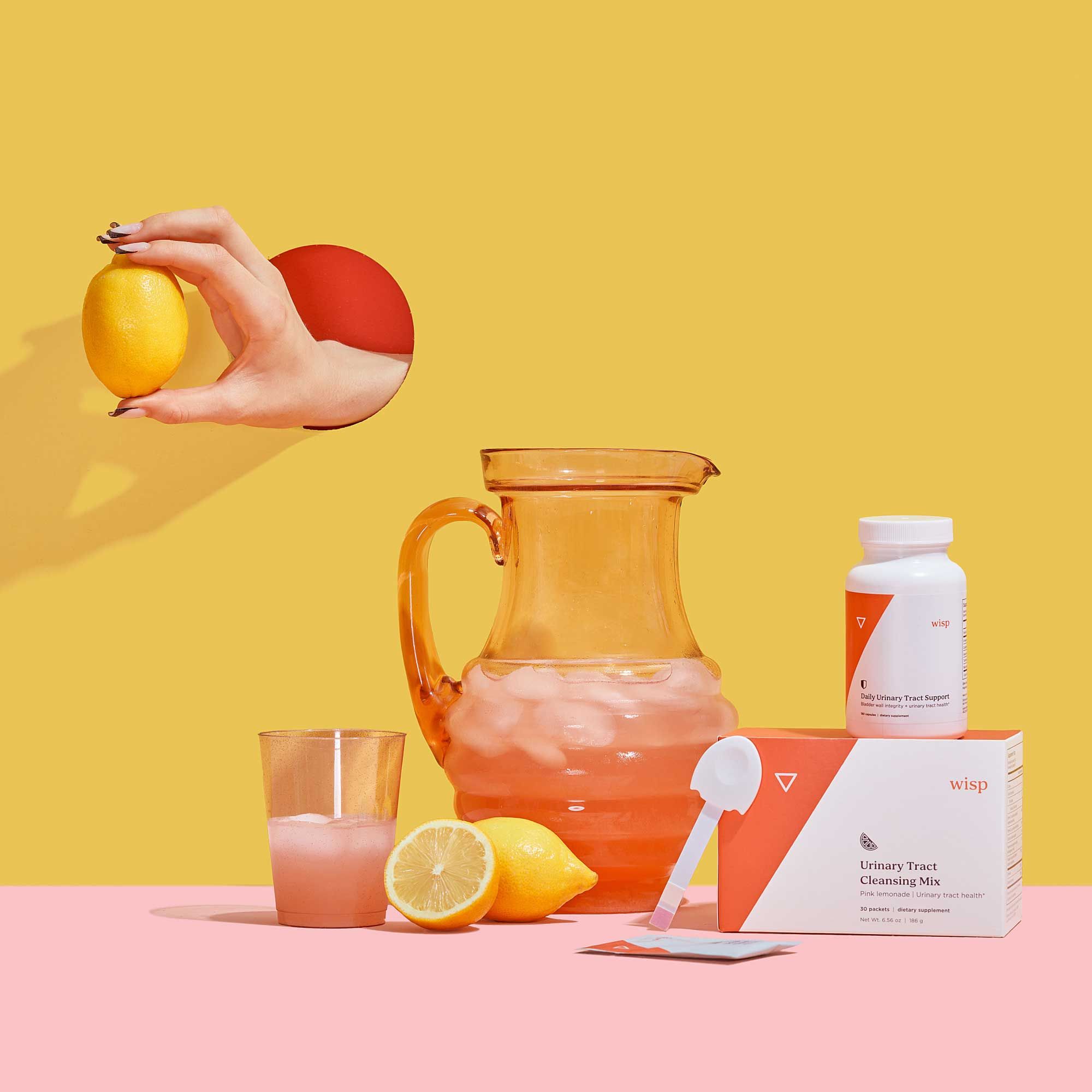 A hand holding a lemon next to a pitcher of pink lemonade with Wisp's Urinary Tract Duo packaging on a pink surface with a yellow background