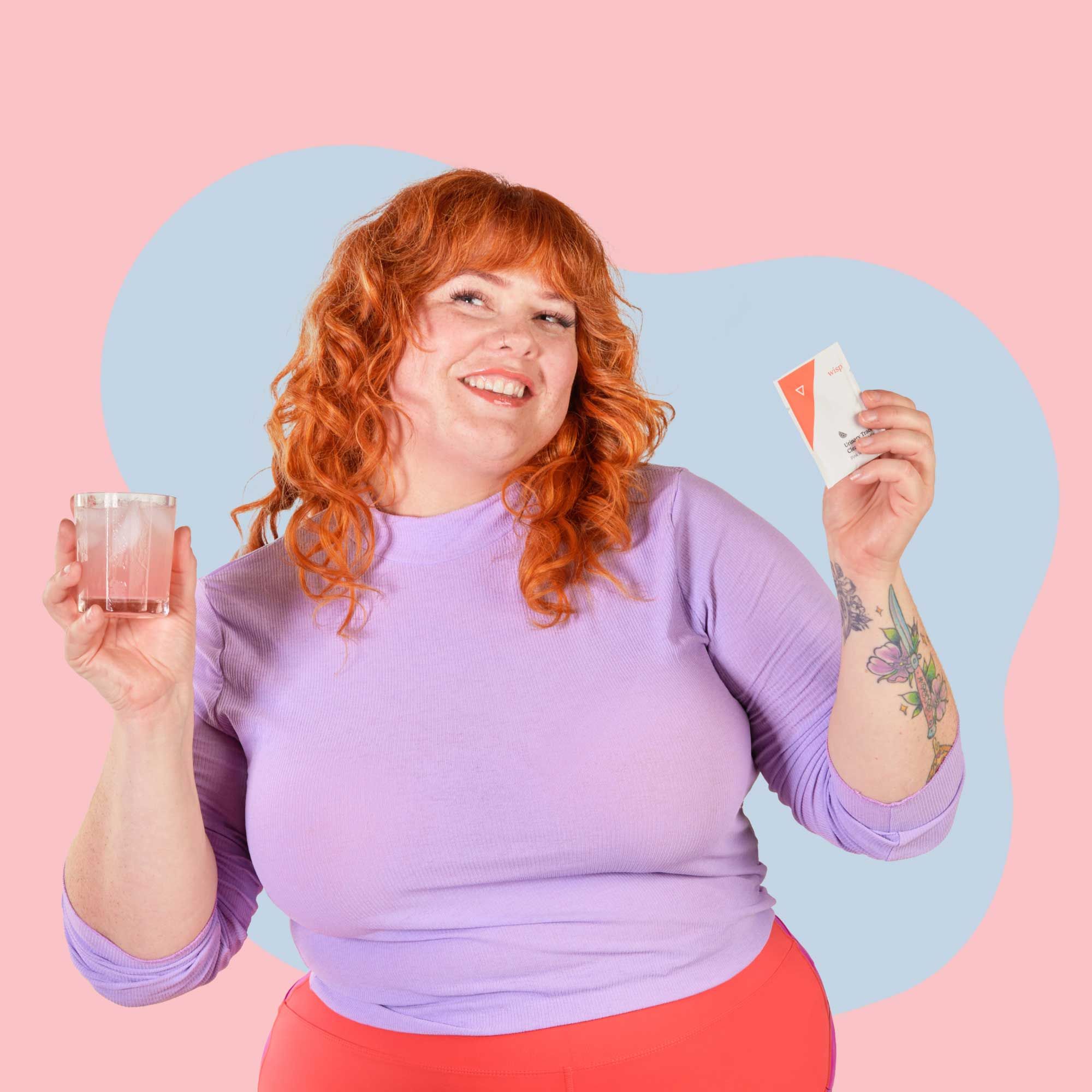 A woman wearing a purple top is holding a drinking glass and a packet of Wisp Urinary Tract Cleansing Mix while looking happy