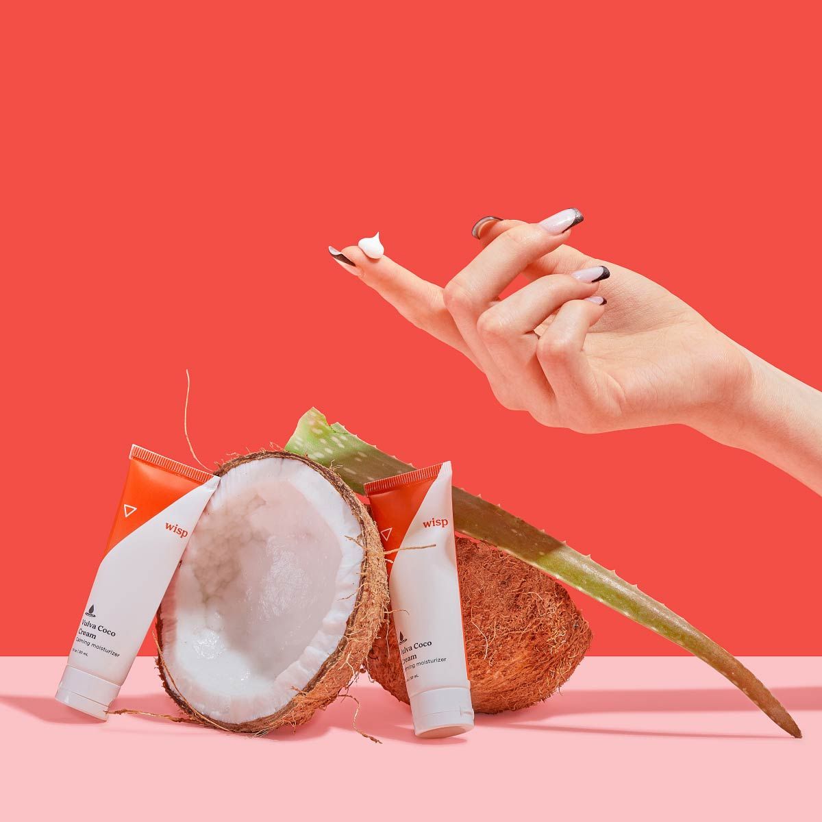 A woman's hand with a dollop of Wisp Vulva Coco Cream on her finger and two bottles of the cream next to an opened coconut and an aloe very leaf on a pink surface with a red background