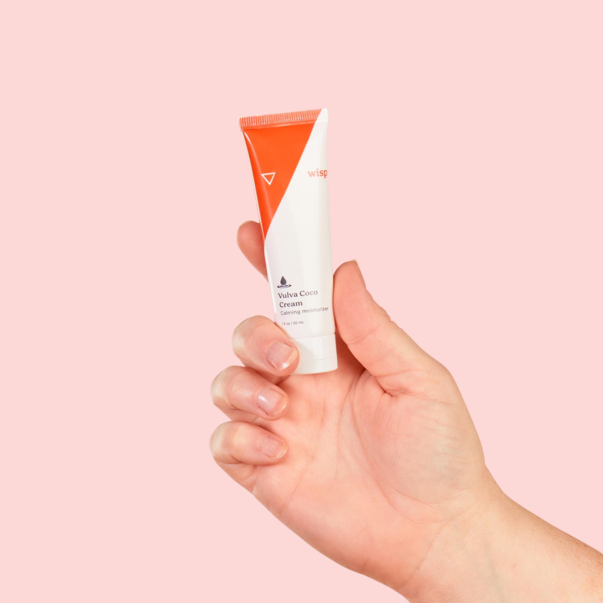 A woman's hand holding Wisp Vulva Coco Cream in front of a pink background