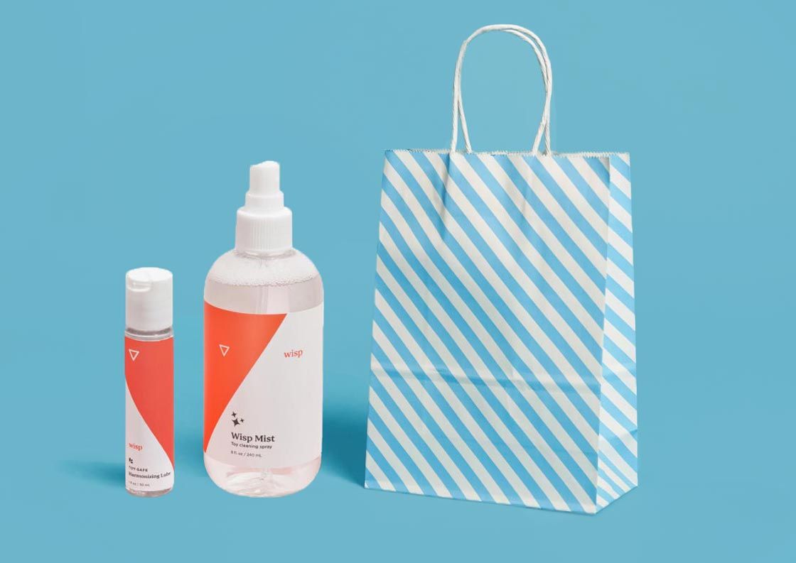 A striped blue and white gifting bag next to Wisp Mist and Wisp Harmonizing Lube on a blue surface