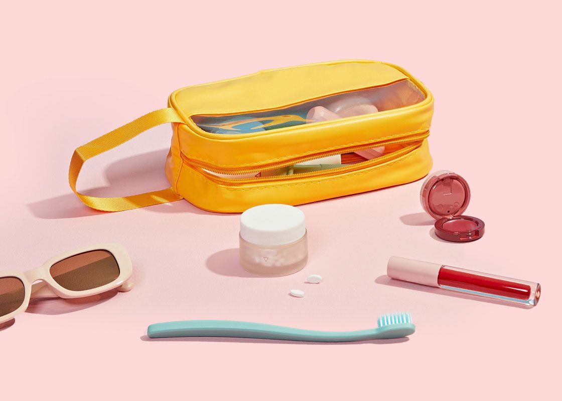 A yellow travel pouch with a Wisp glass pill jar, sunglasses, a toothbrush, lip gloss, and blush on a pink surface