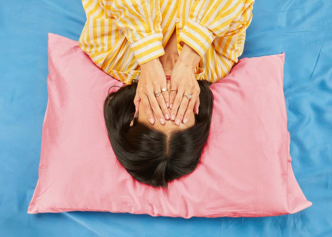 A woman wearing striped yellow and white pajamas laying on a bed with a pink pillow and blue sheets putting her hands over her face