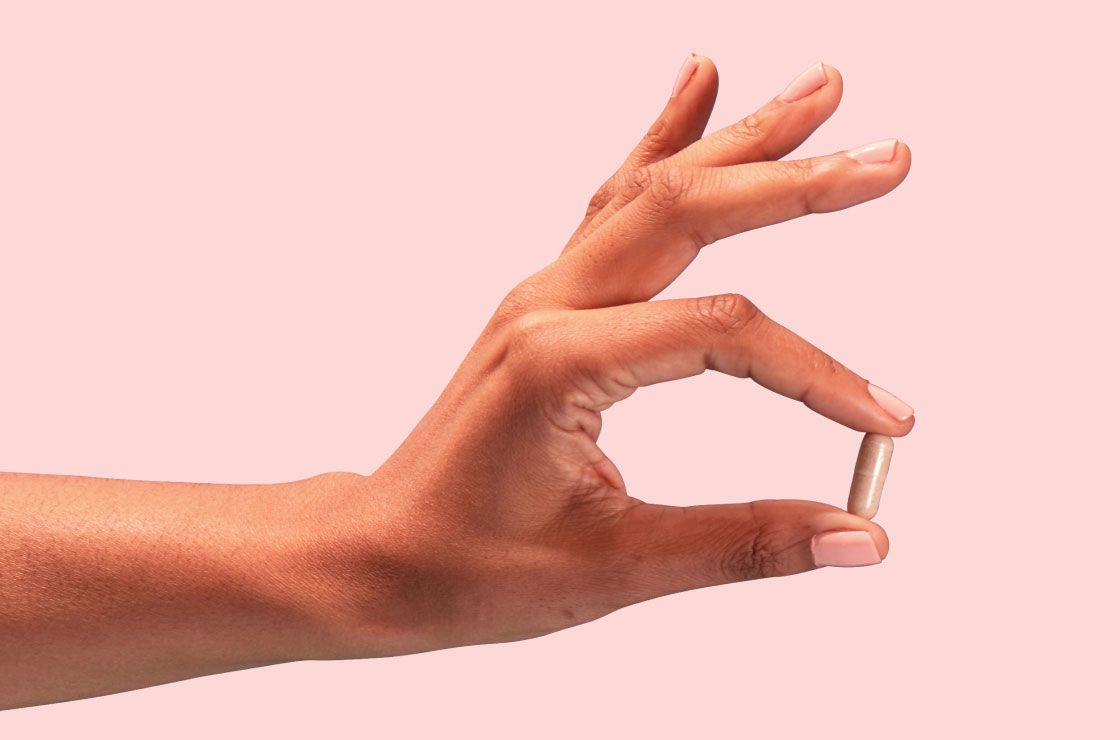 A woman's hand holding a pill in front of a pink background