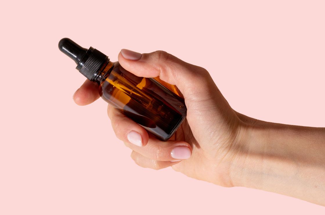 A woman's hand holding an amber dropper bottle in front of a pink background