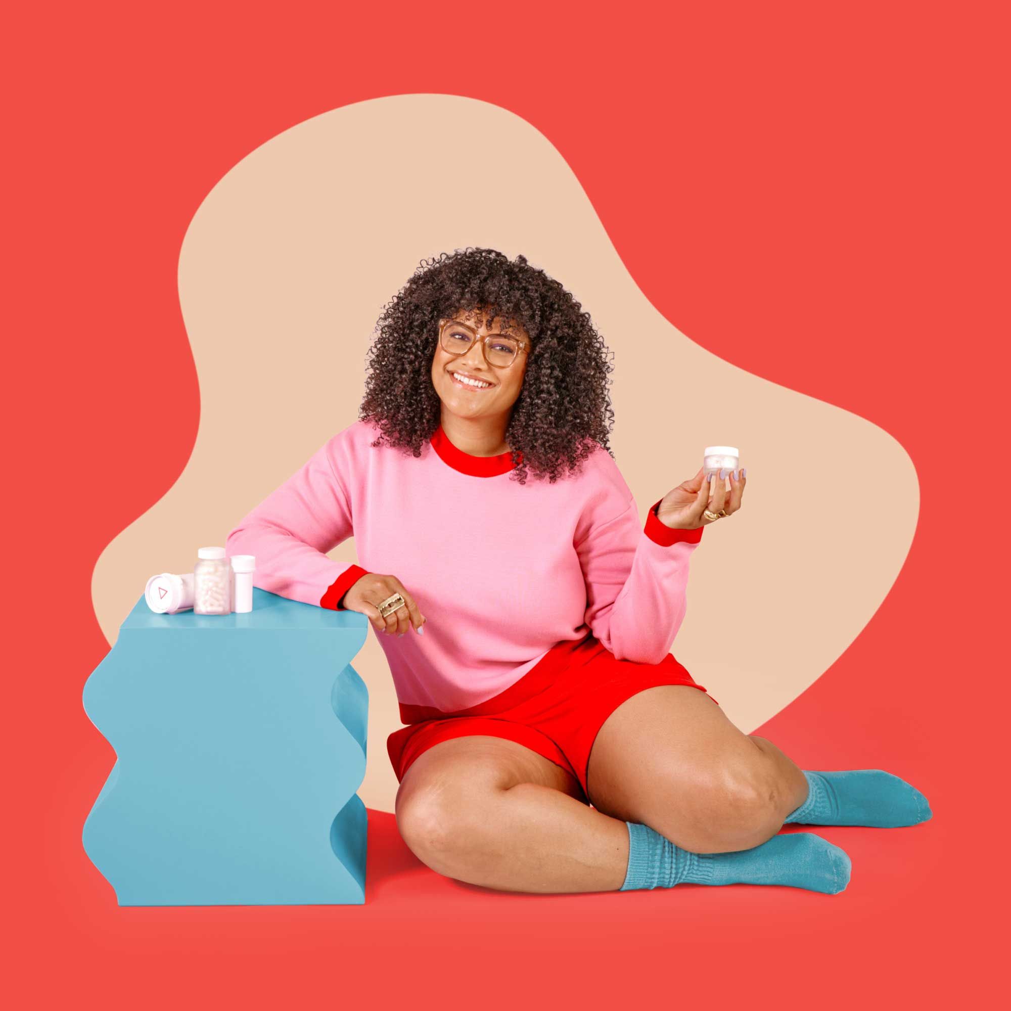 A woman wearing a pink shirt, red shorts, and teal socks is sitting on a red surface next to a teal nightstand with various Wisp products on it