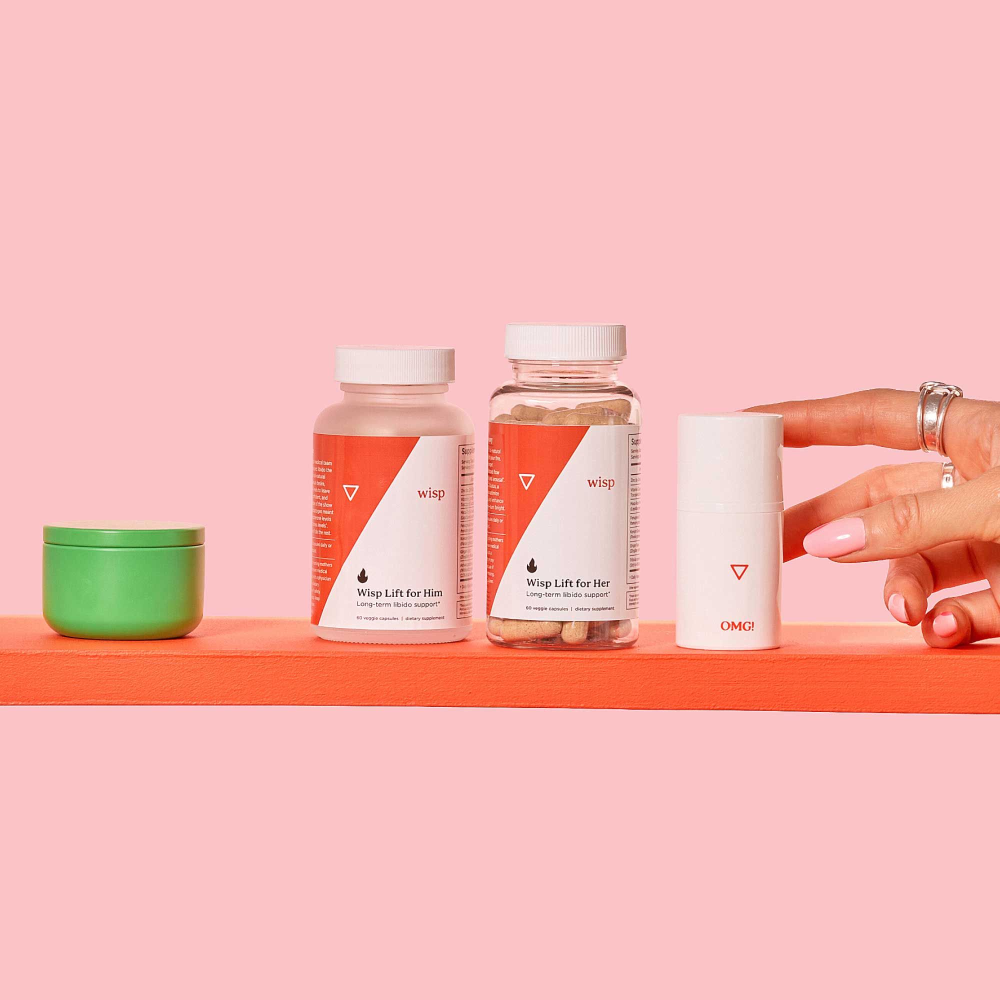 A woman's hand reaching for OMG Cream next to bottles of Wisp Lift for Her and Wisp Lift for Him on a red shelf with a pink background