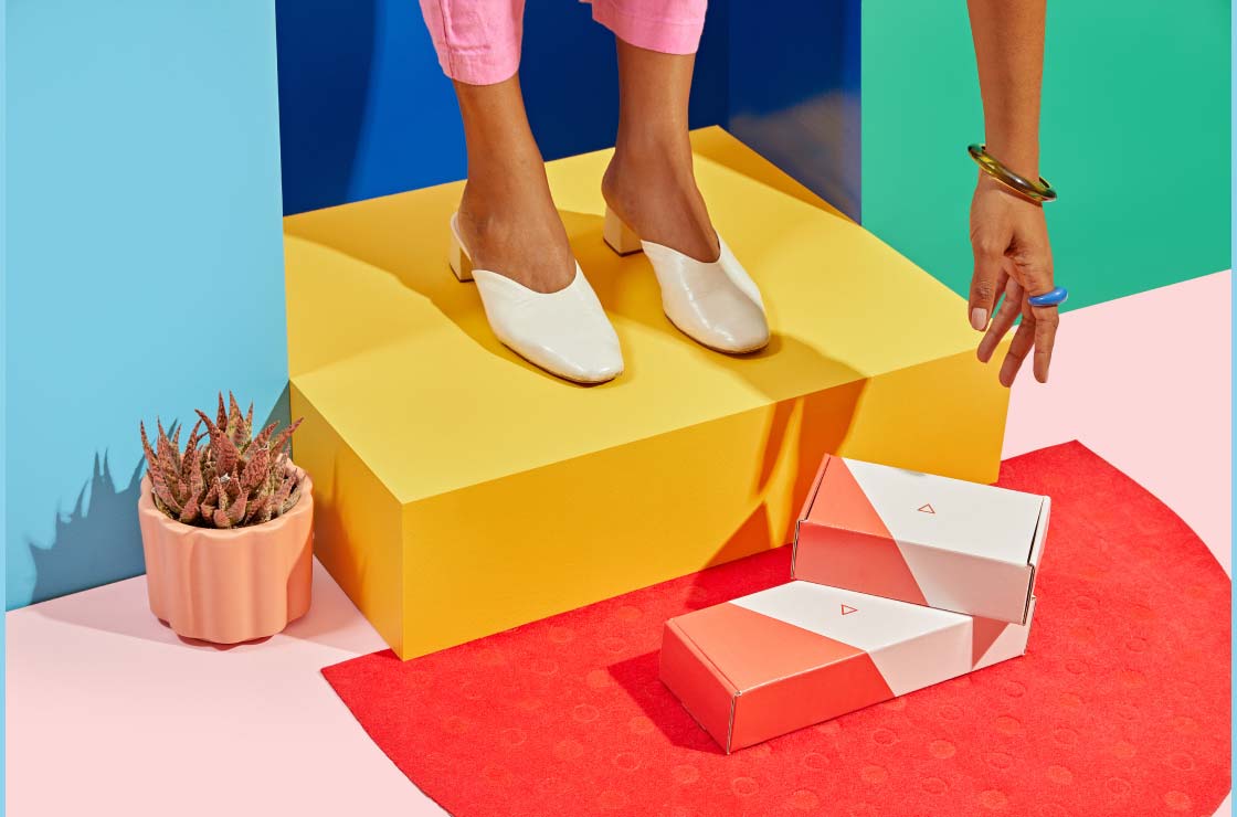 A woman standing on a porch is reaching for Wisp shipping boxes on a red door mat, a pink floor and blue and green walls