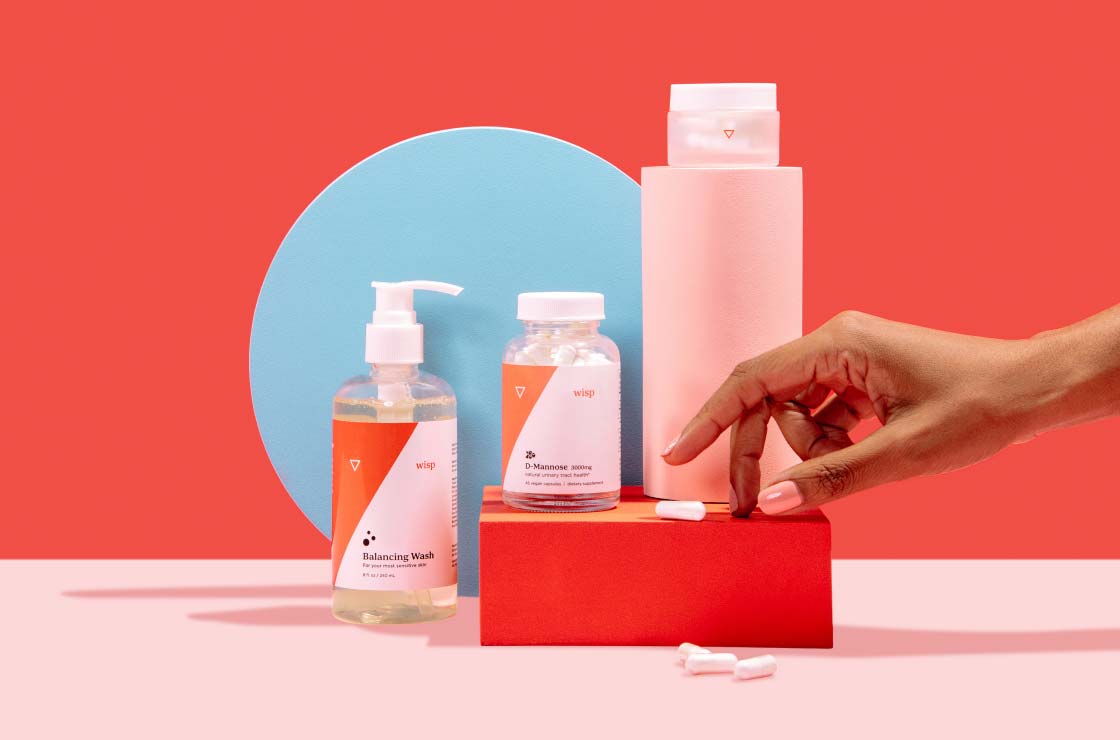 A woman's hand reaching for a pill on a red box next to various Wisp products balanced on colorful abstract shapes on a pink surface with a red background
