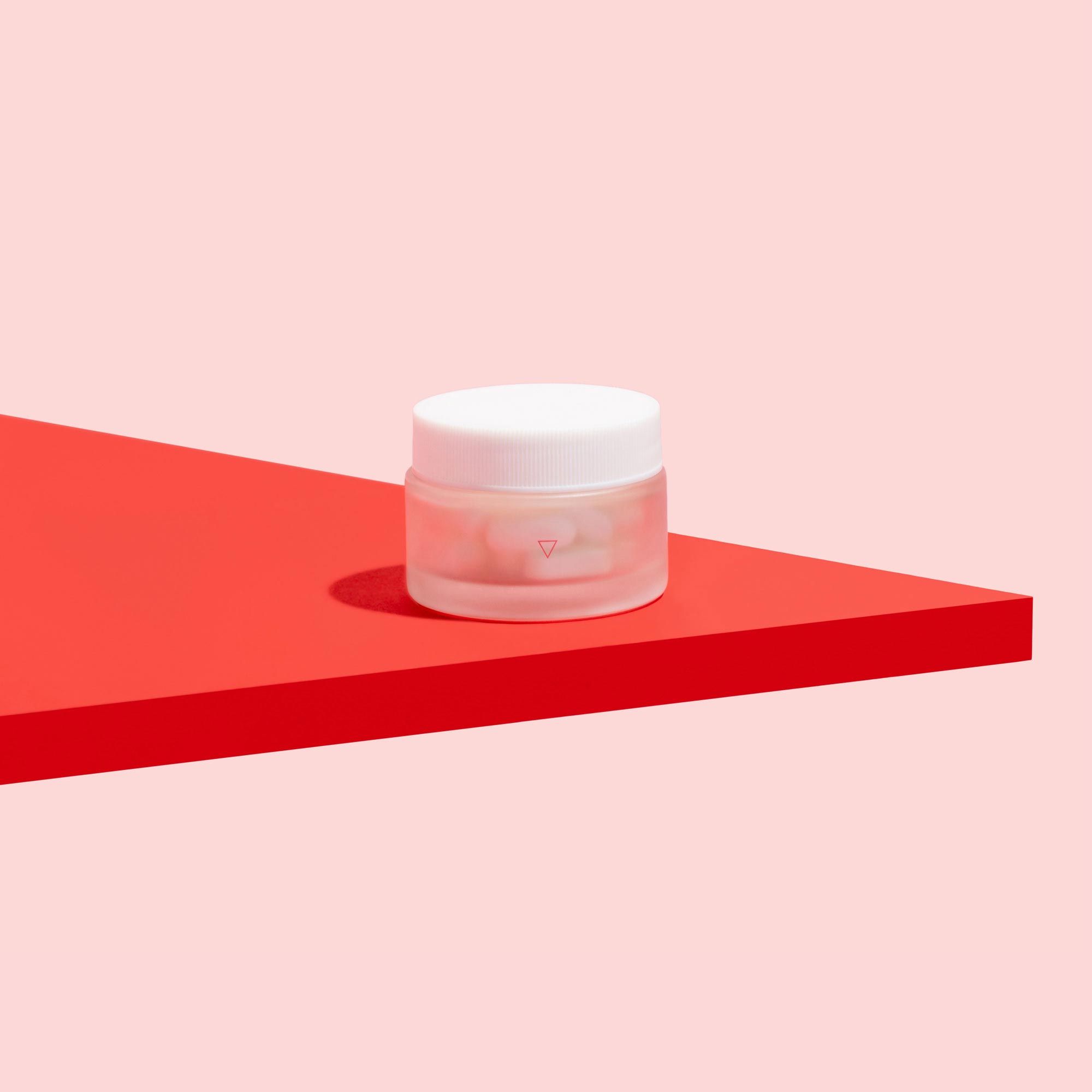 Wisp small glass pill jar on a red surface with a pink background