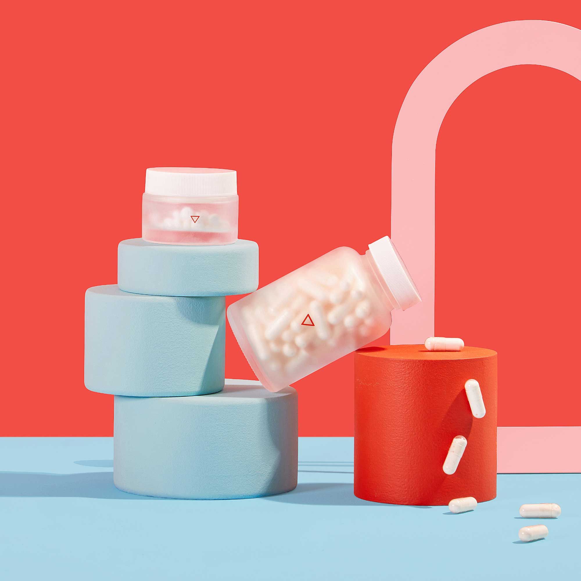A small and large Wisp glass pill jar balanced on colorful abstract shapes with pills spilling out on a blue surface with a red background