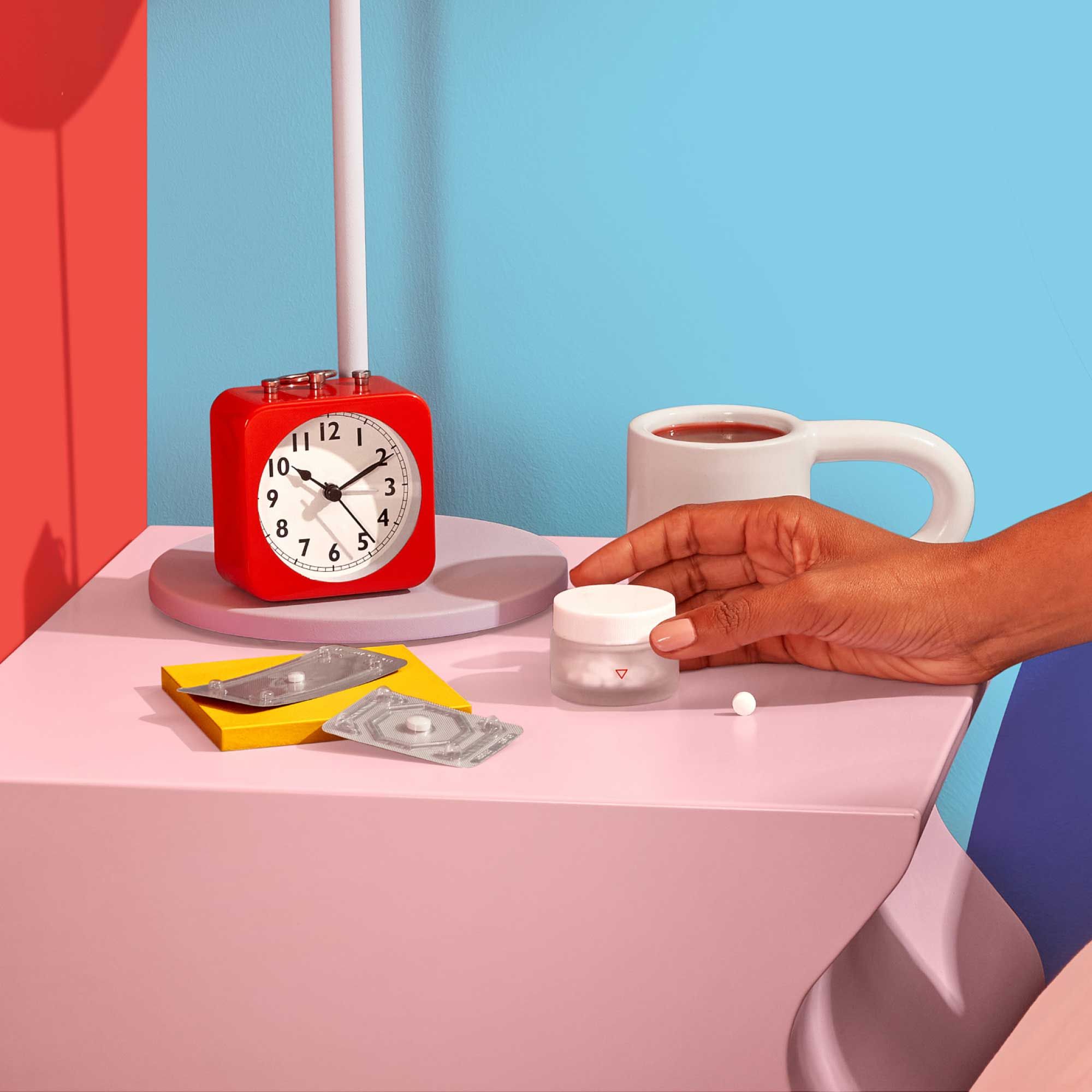 A woman's hand reaching for a Wisp small glass pill jar on a pink nightstand with emergency contraception packets, a red alarm clock and a mug with a light blue background