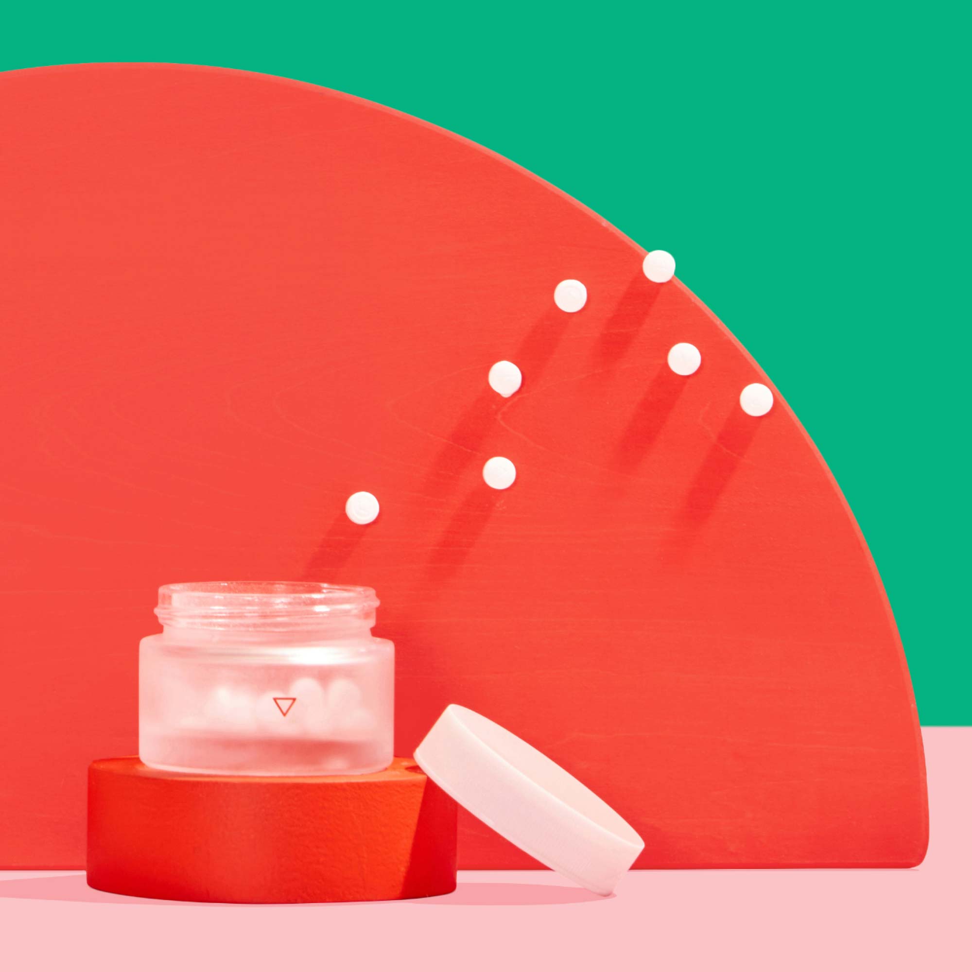 A Wisp small glass pill jar with pills floating out of it near colorful abstract shapes on a pink surface with a green background