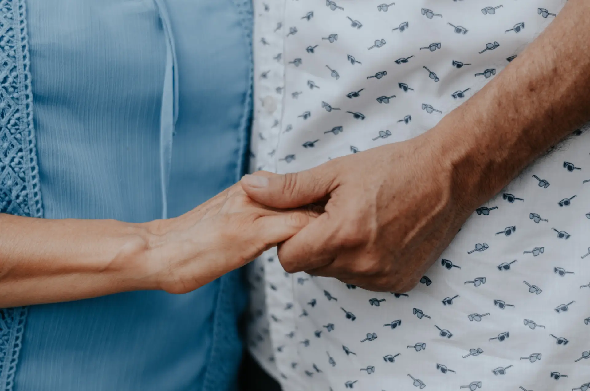 older woman wearing a blue top and older man wearing a white and black polka dot shirt holding hands