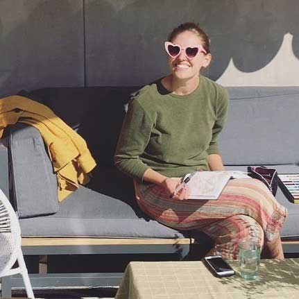 Artist Allie Sullberg wearing pink heart sunglasses, a green sweater and striped pants sitting on a sofa outside holding a notepad