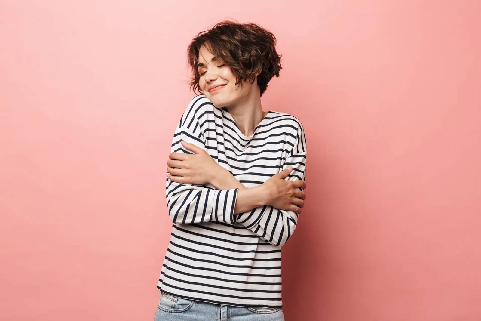 woman in striped shirt hugging herself against a pink background