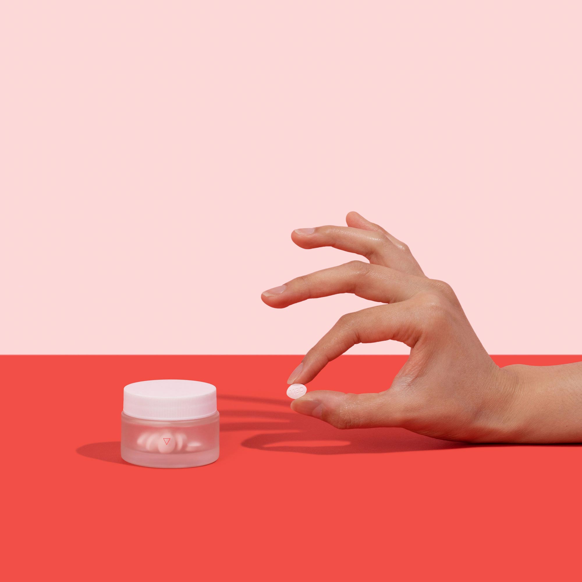 Woman's hand holding pill to treat yeast infections next to jar of antifungals on a red surface, on a pink background