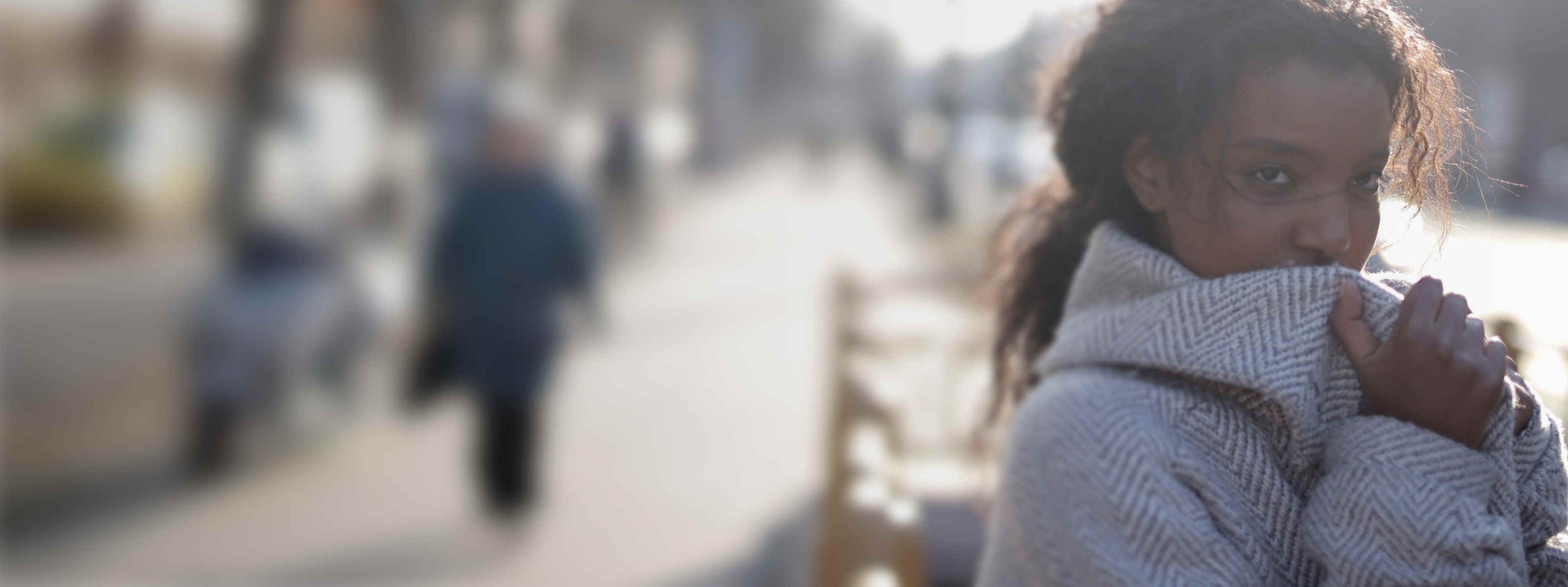 Woman bundled in a scarf and coat, pulling the fabric up over her chin against the cold on a blurred city sidewalk.