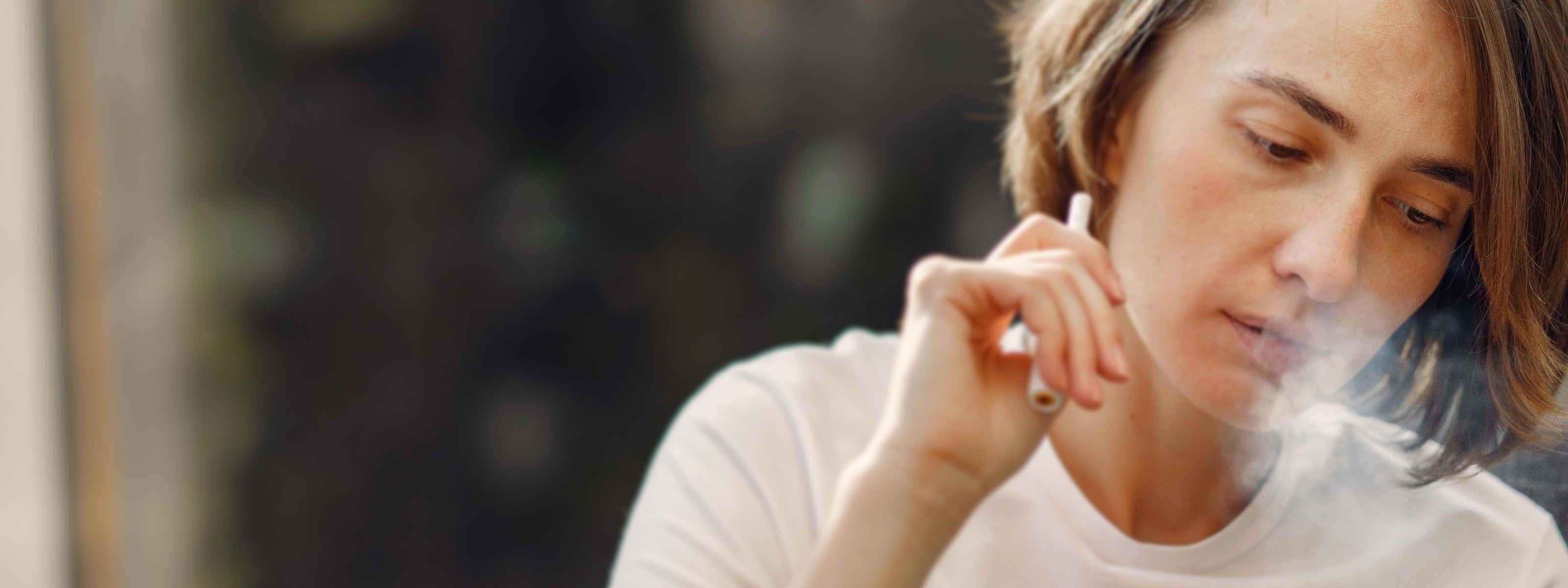 A woman in a white T-shirt sits holding a vape pen, exhaling a cloud of vapor as she looks downward.