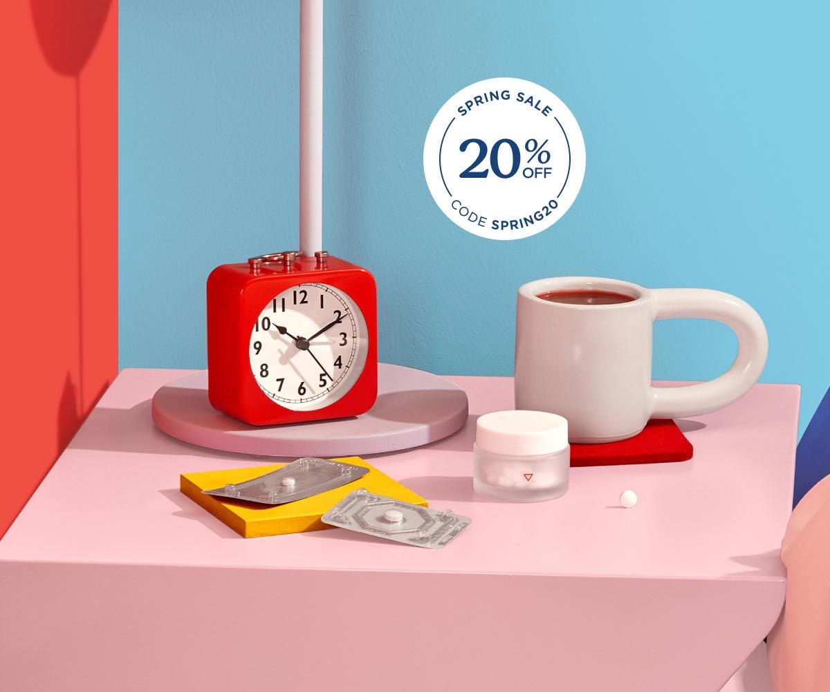 A Wisp glass pill jar and 2 emergency contraception packets are on a pink nightstand with a red alarm clock and white mug next to a bed with colorful bedding