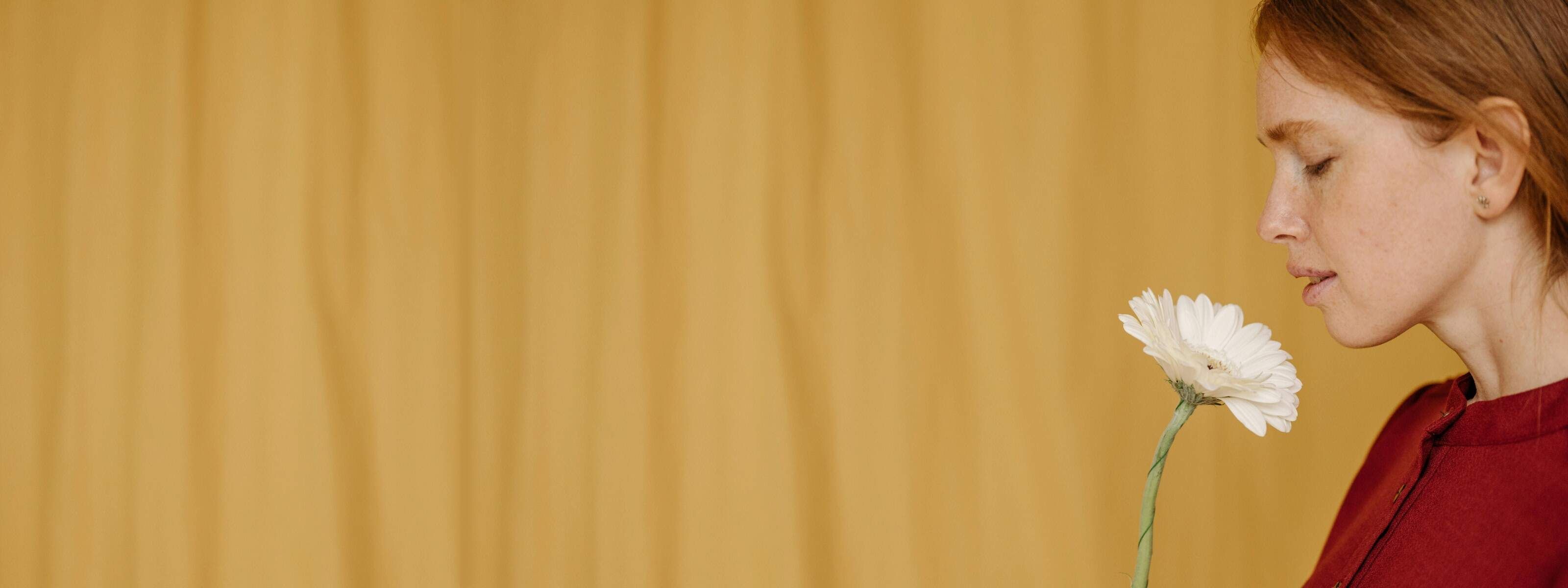 A person with red hair smelling a white flower against a yellow background