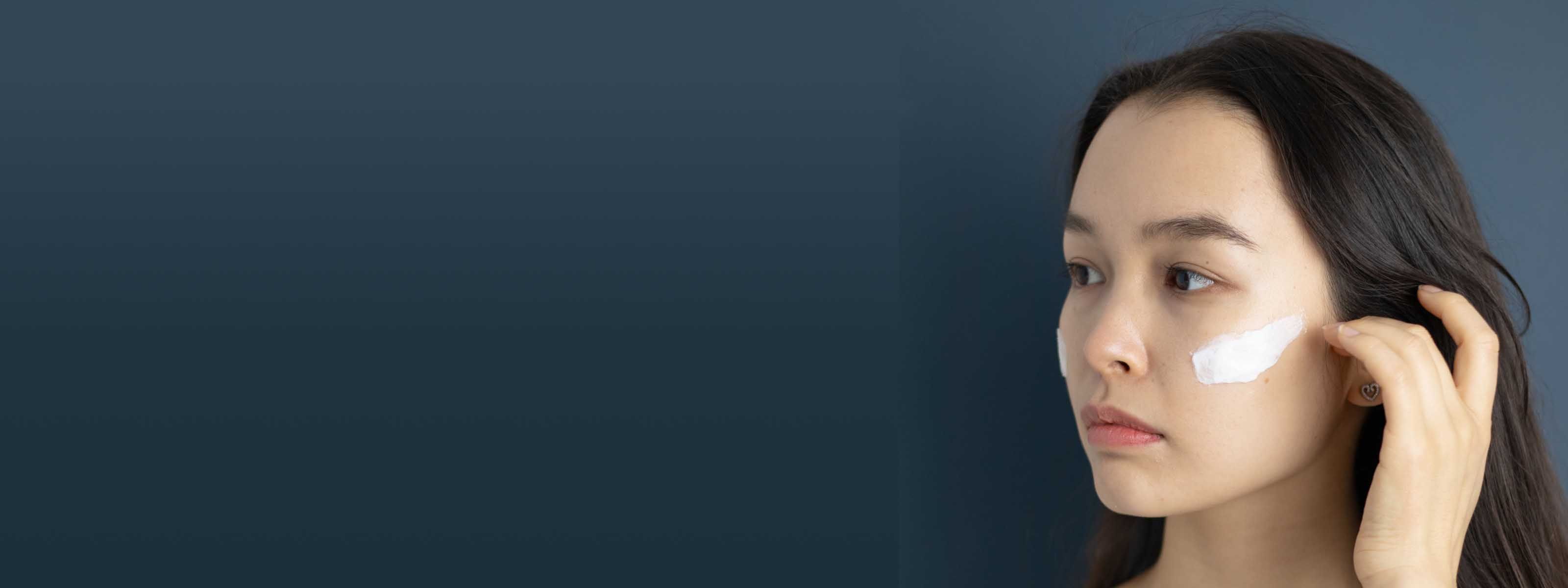 A pretty woman with dark hair has a swipe of cream on her cheek in front of a dark blue background